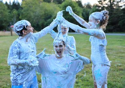 Campers and a counselor, covered in shaving cream, laugh as they make funny shapes with their hair covered in colored foam.